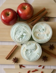 Three containers of a green and white sugar scrub on a wooden board with apples, cinnamon sticks, and star anise.
