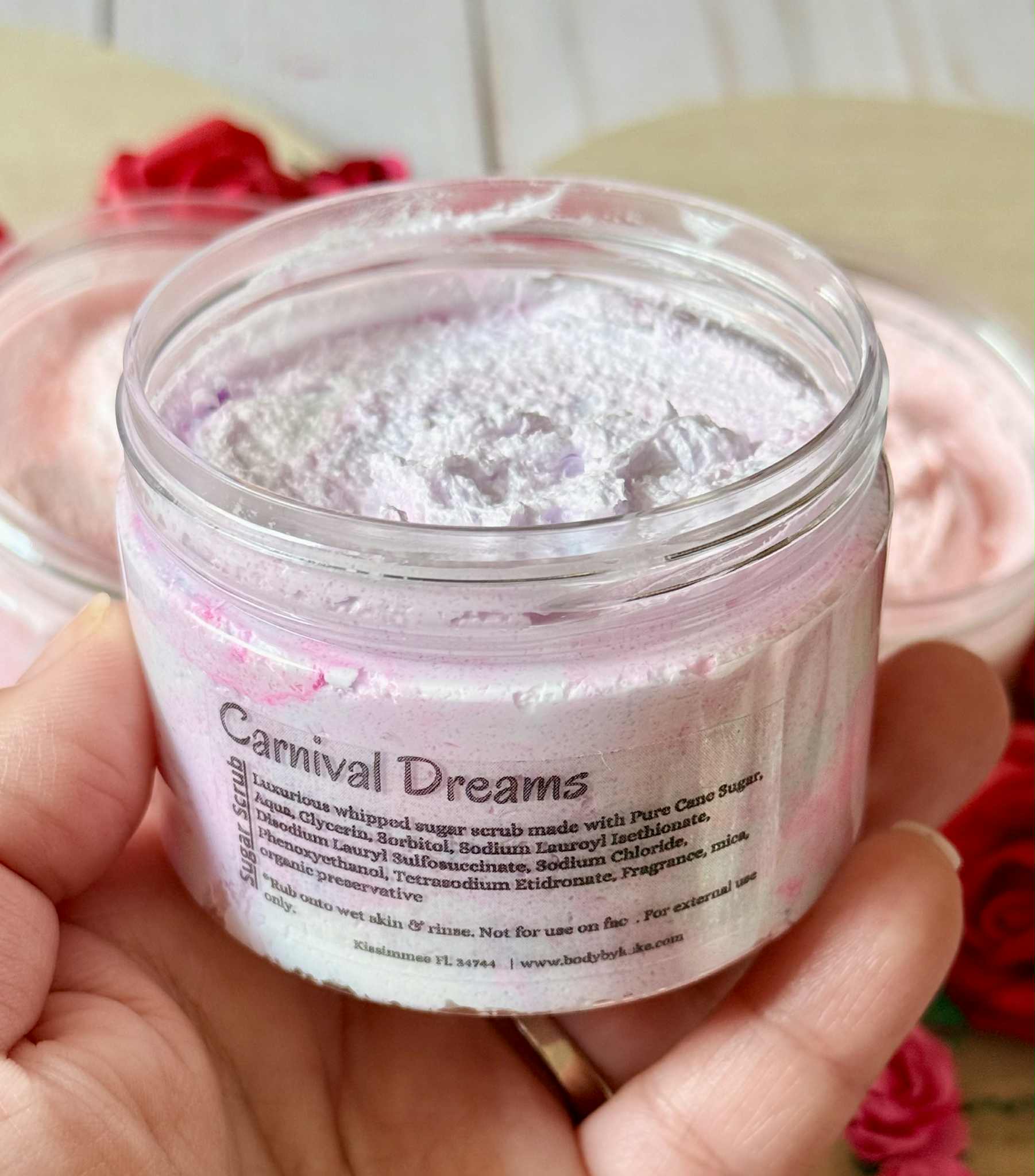 Jar of 'Carnival Dreams' whipped sugar scrub held by a hand with flowers in the background
