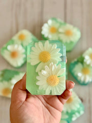Hand holding a green soap bar with daisy flowers against a neutral background