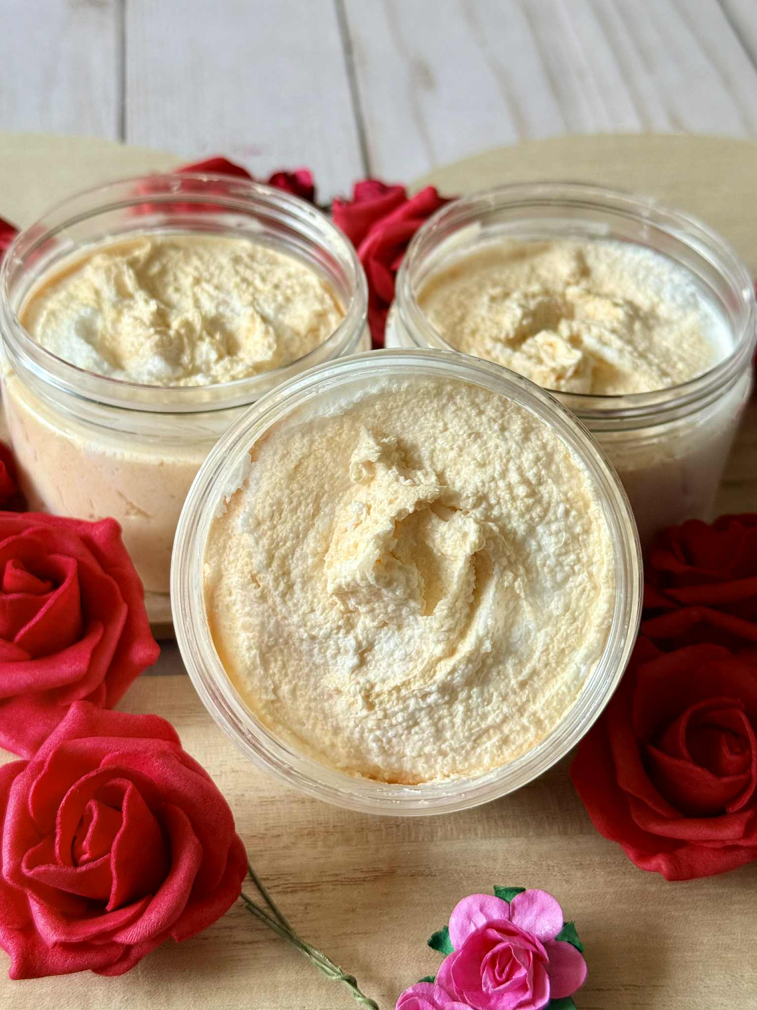 Three jars of orange sugar scrub on a wooden surface with red and pink flowers.