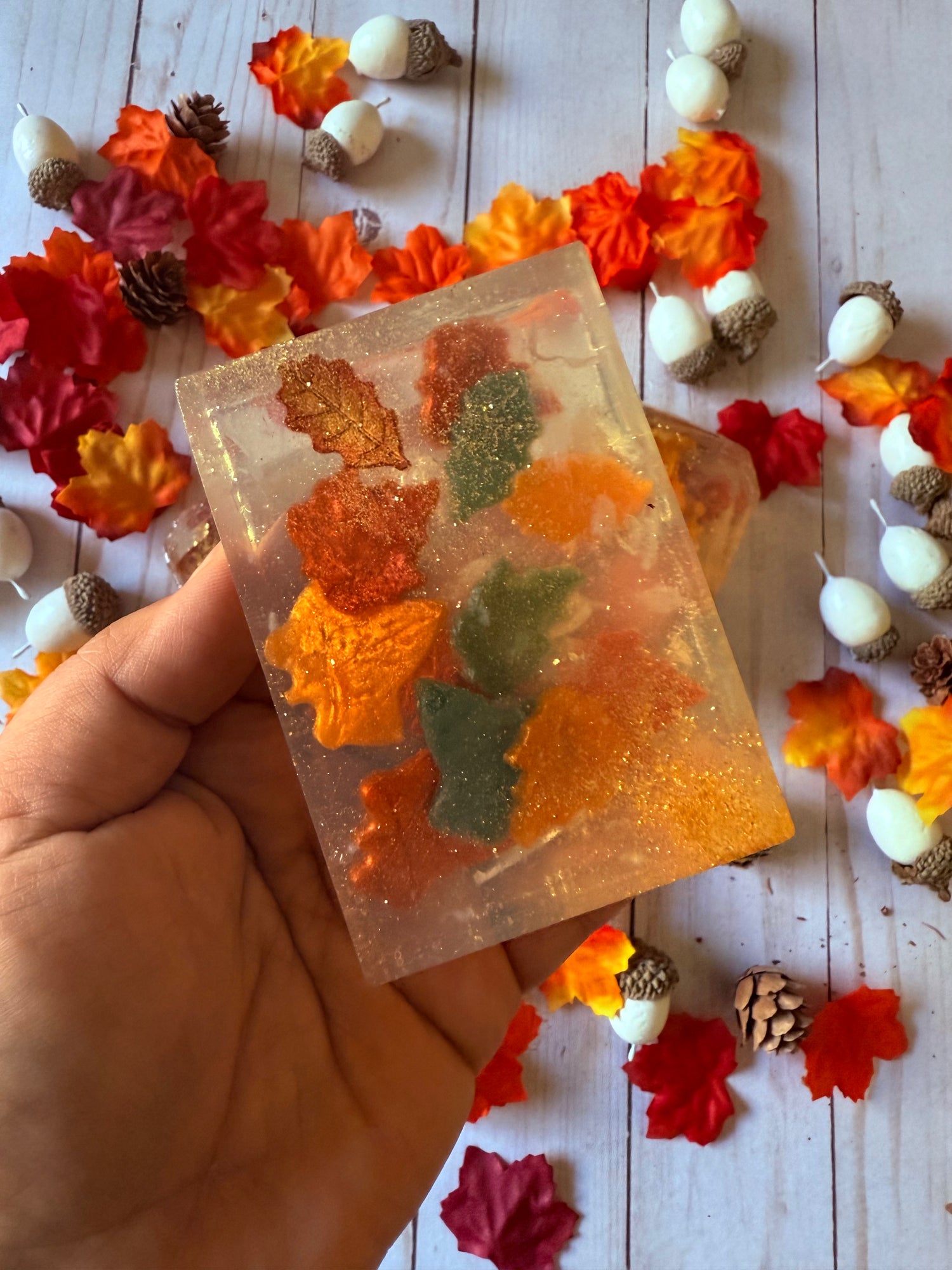 Hand holding a soap bar with autumn leaf decorations on a wooden surface with fall-themed decor.