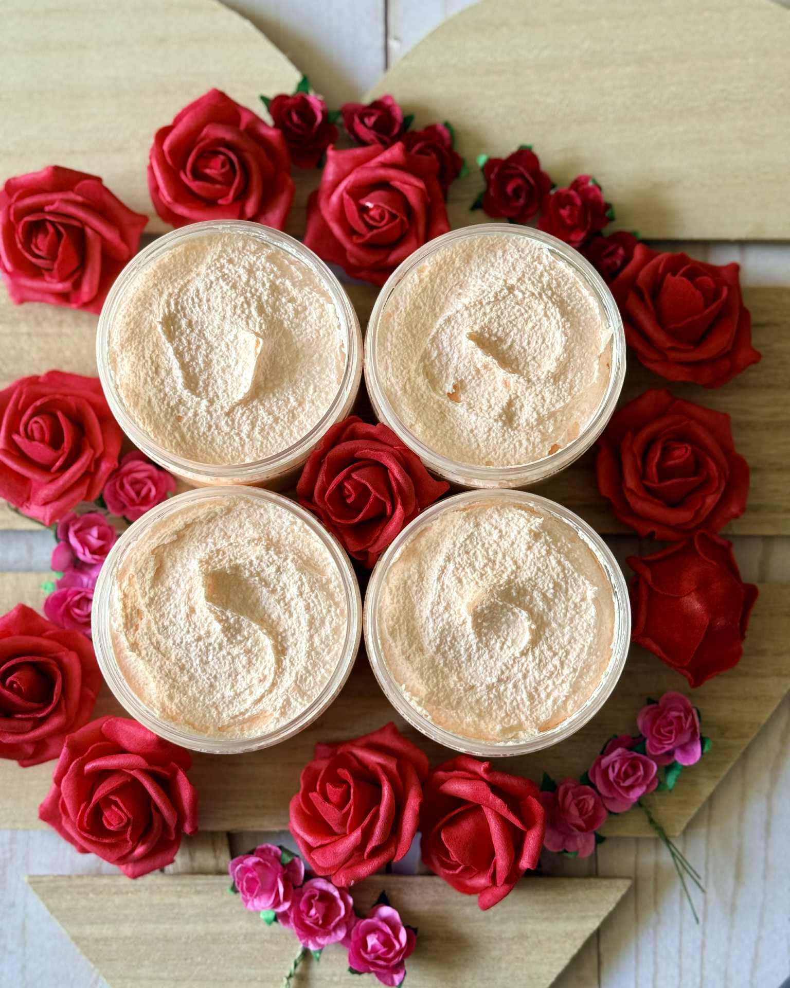 Four containers of a beige product surrounded by red and pink roses on a wooden heart-shaped board.