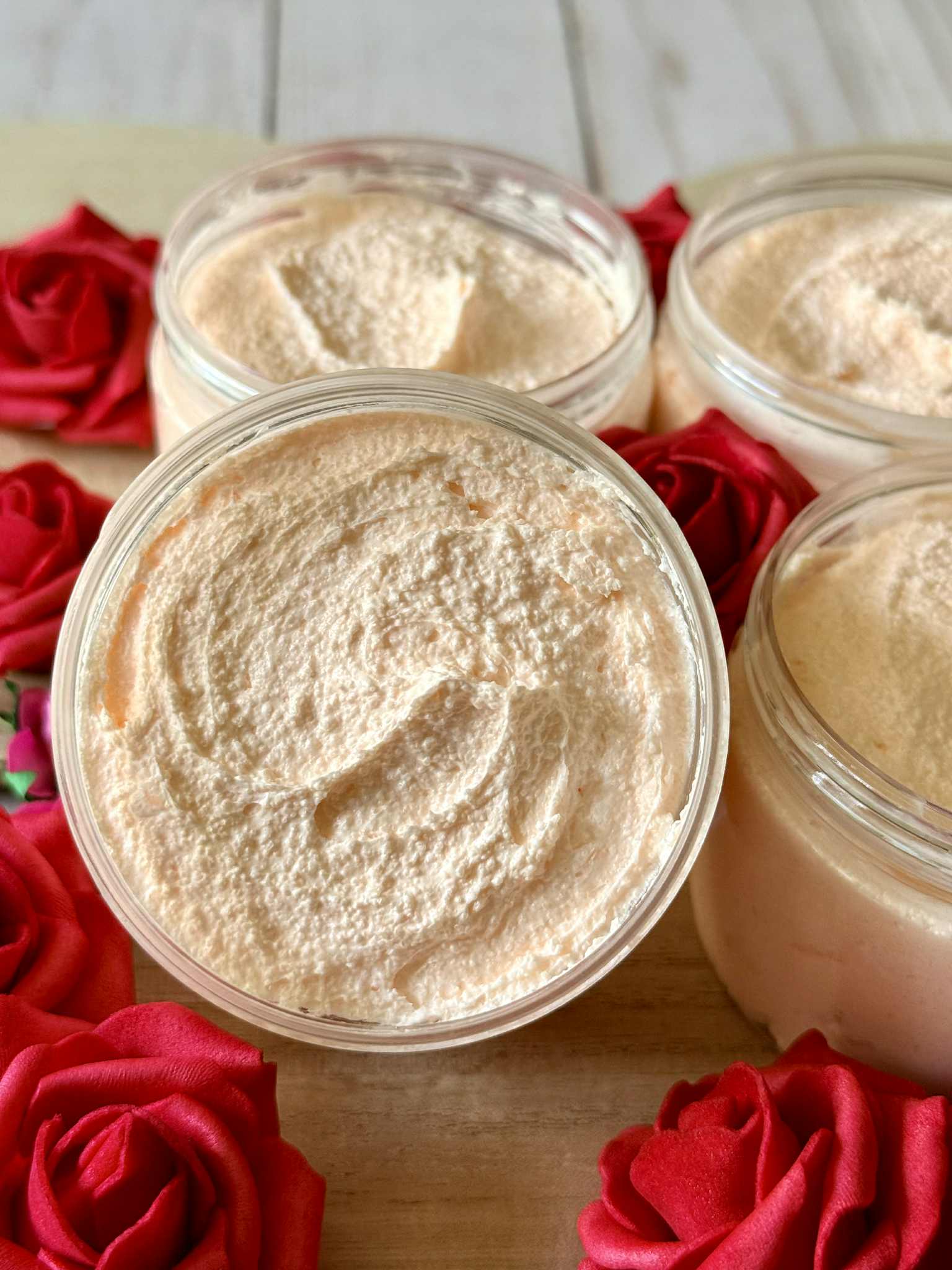 Four containers of beige cream on a wooden surface with red roses around them