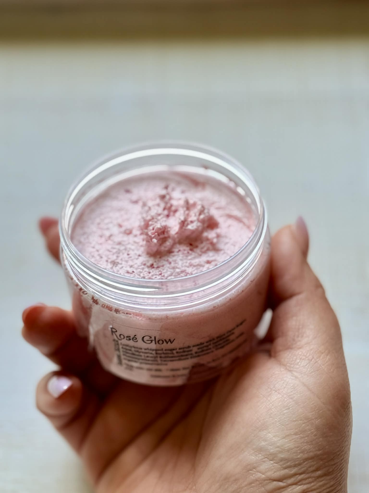 Hand holding a jar of pink 'Rose Glow' scrub against a neutral background