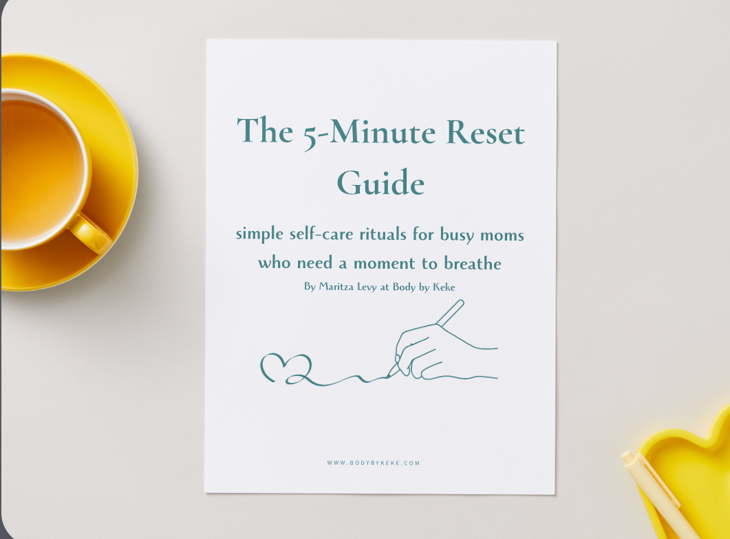 A white booklet or guide titled "The 5-Minute Reset Guide" lies on a light neutral surface, styled with a yellow cup on the left and a yellow heart-shaped dish on the right. The cover has teal text and a simple line drawing of a hand writing a heart.