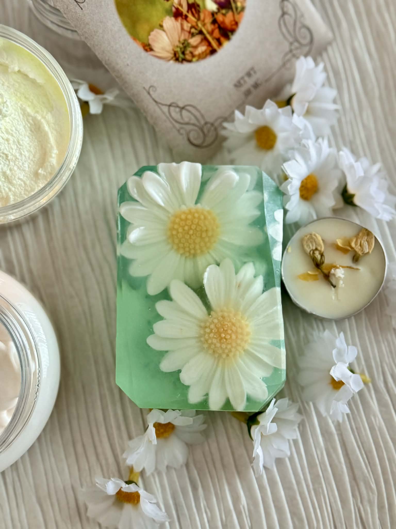 Handmade soap with daisy design on a wooden surface with flowers, tea light candle and containers.