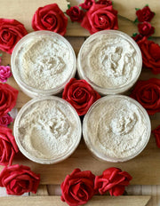 Four containers of white sugar scrub surrounded by red roses on a wooden surface.