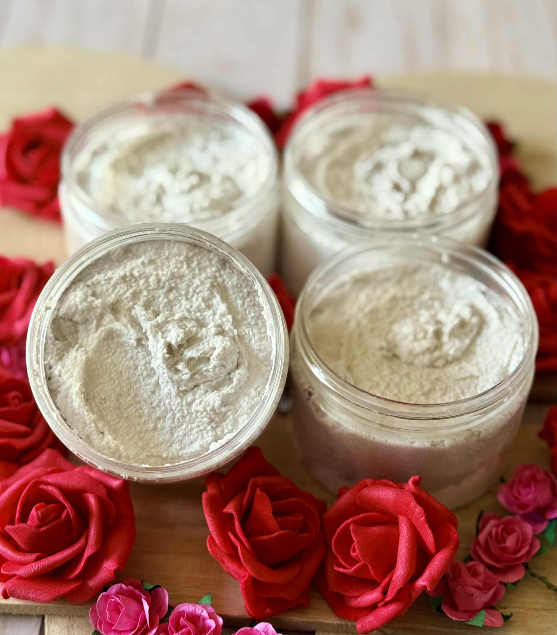 Four jars filled with white sugar scrub on a wooden board surrounded by red and pink roses.
